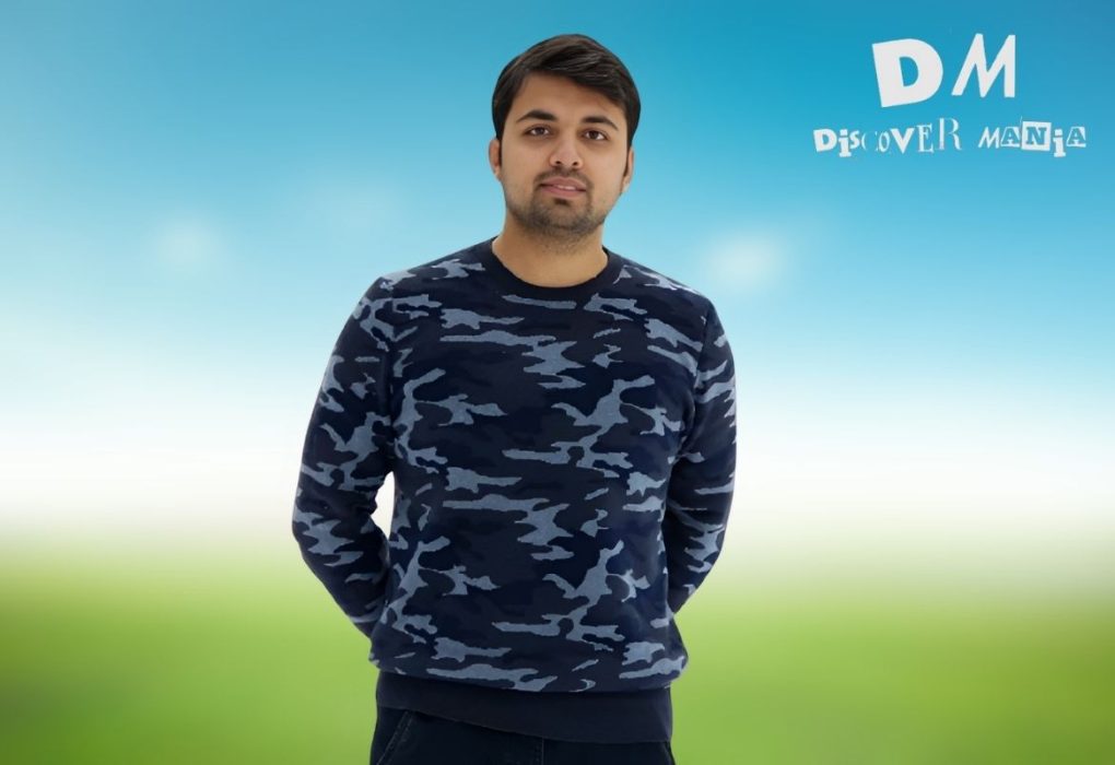 Manish Kumar Shukla Seattle standing in front of a blue sky background with a blurred green field, wearing a camouflage-patterned sweatshirt. The logo "DM Discover Mania" appears in the top right corner.